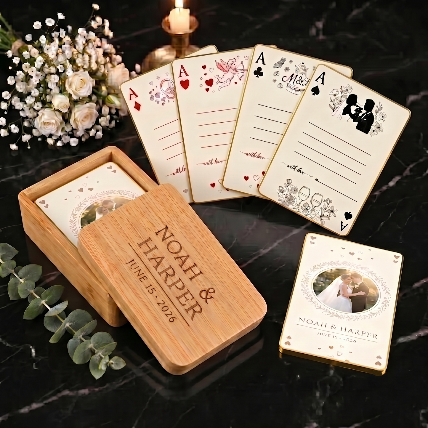 Custom wedding playing cards in an engraved bamboo wood box with gold-edged cards and four signature designs on black marble with white roses and eucalyptus
